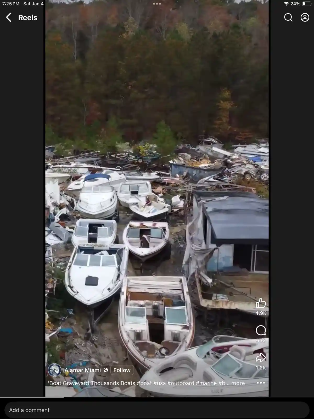 An aerial view shows a large, muddy lot filled with hundreds of abandoned, decaying boats and boat parts, surrounded by…