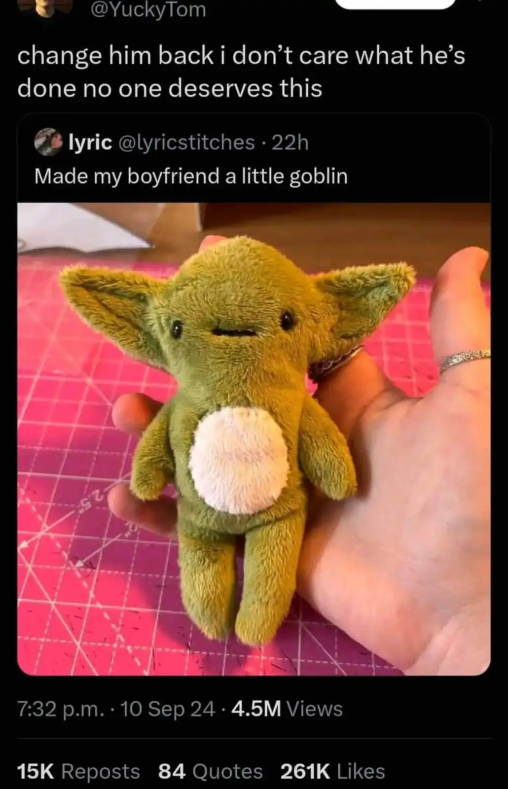 A hand holds a small, green, plush toy resembling a goblin with large ears and a white belly patch, set against a pink…