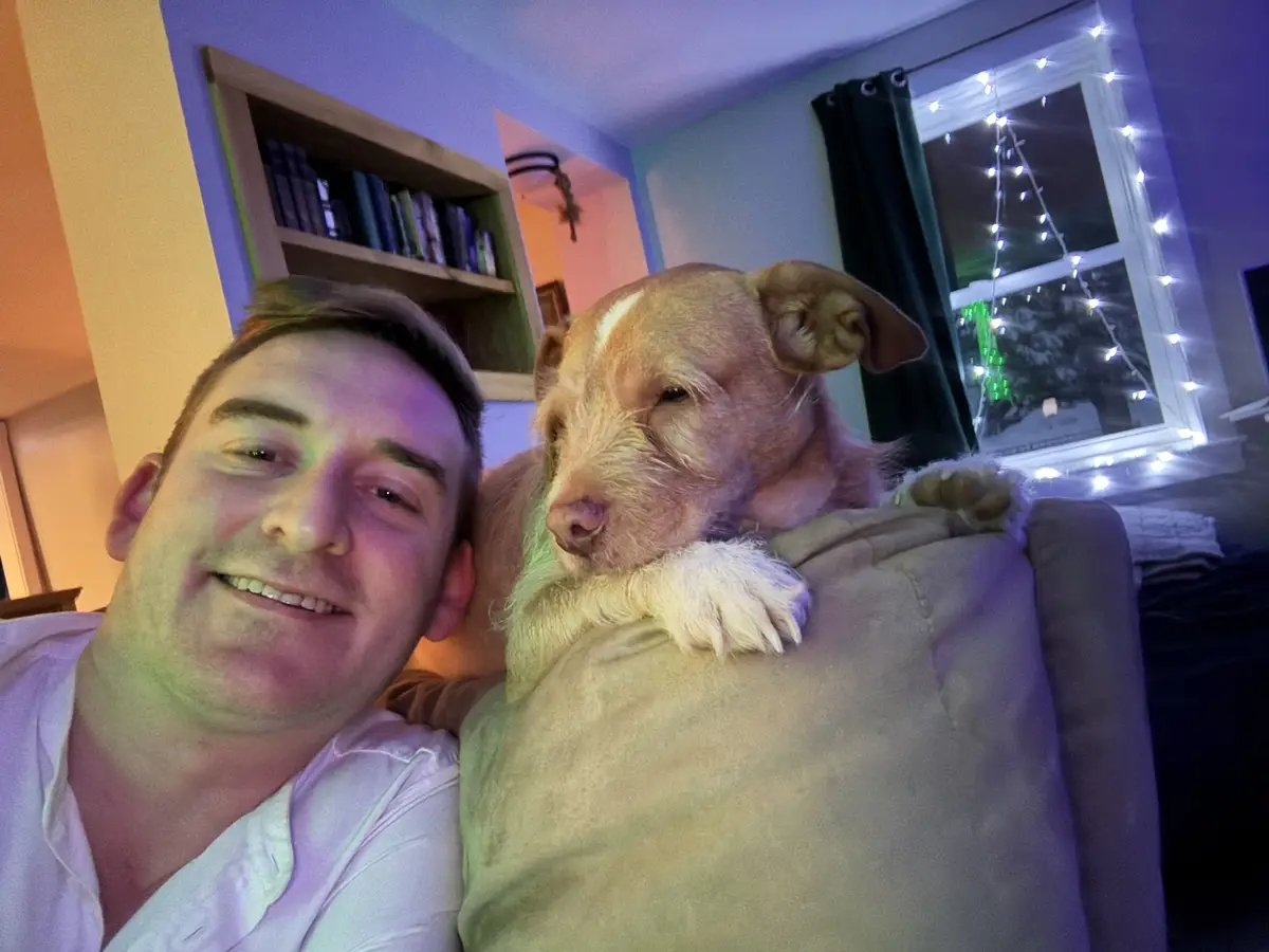 A man in a white shirt smiles at the camera while sitting on a couch next to a light brown dog with one paw resting on the…