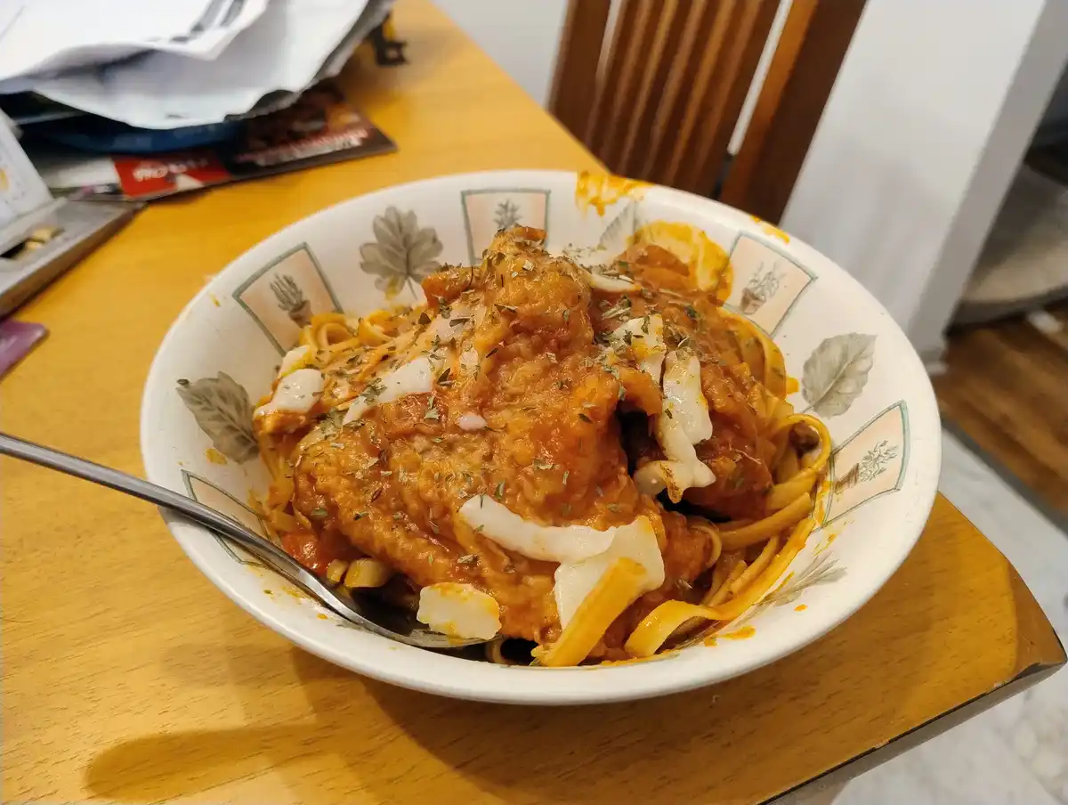 A close-up photo shows a bowl of pasta covered in a thick orange sauce, topped with what appears to be fried chicken pieces…