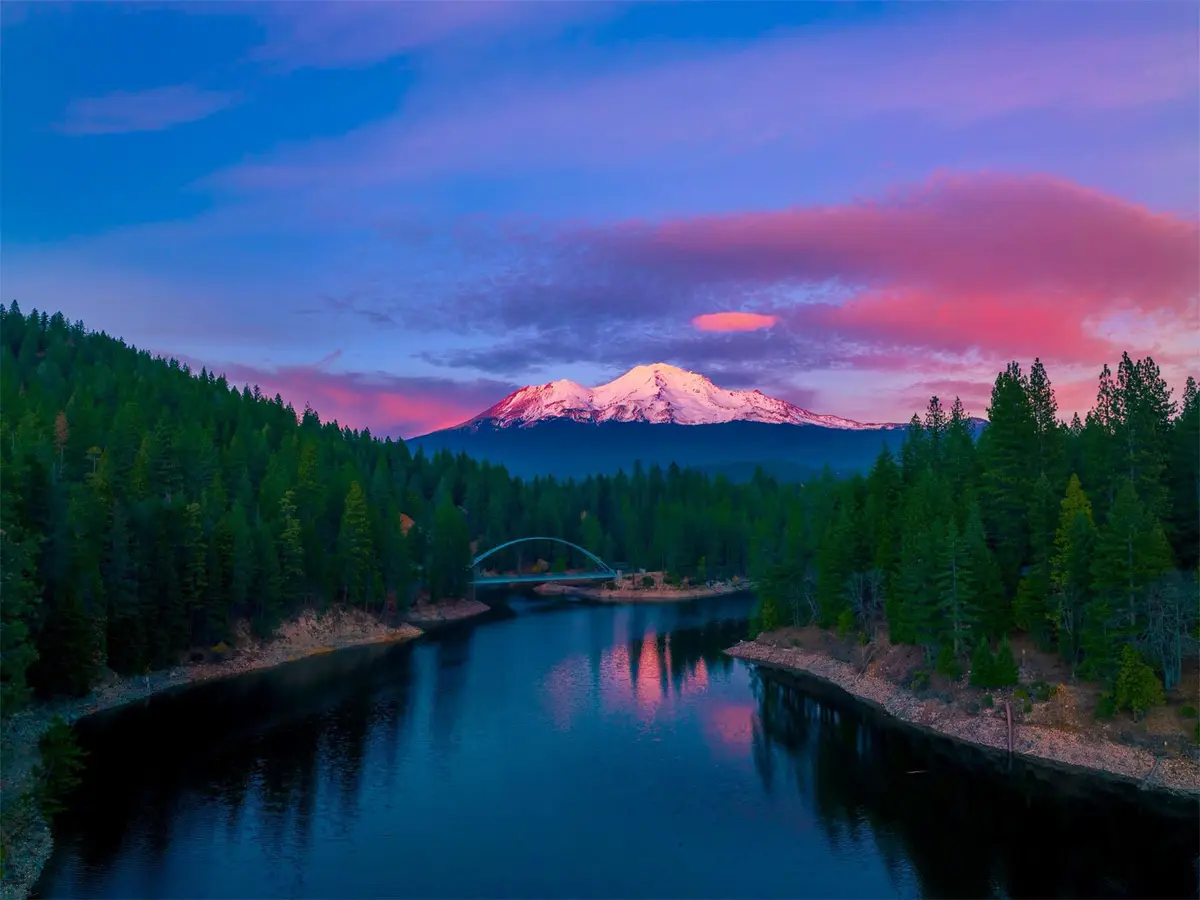 A serene landscape photo shows a snow-capped mountain under a vibrant pink and blue twilight sky, reflected in a calm river…