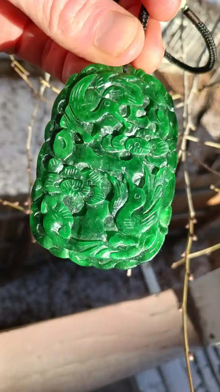 A hand holds a vibrant green, intricately carved jade pendant featuring detailed floral and possibly animal motifs…