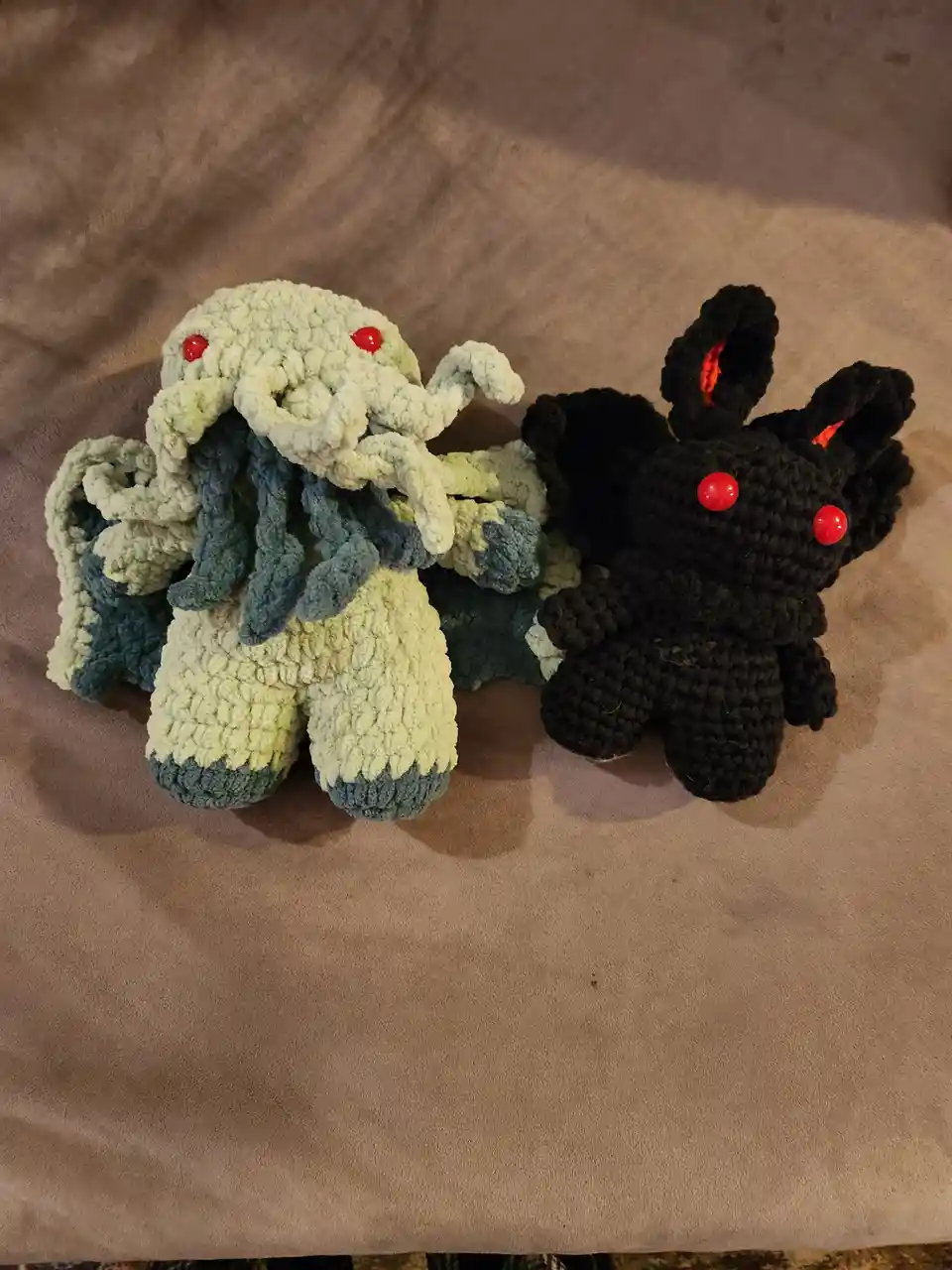 Two handmade crocheted plush toys sit side by side on a brown fabric surface: a pale green Cthulhu with red button eyes and…