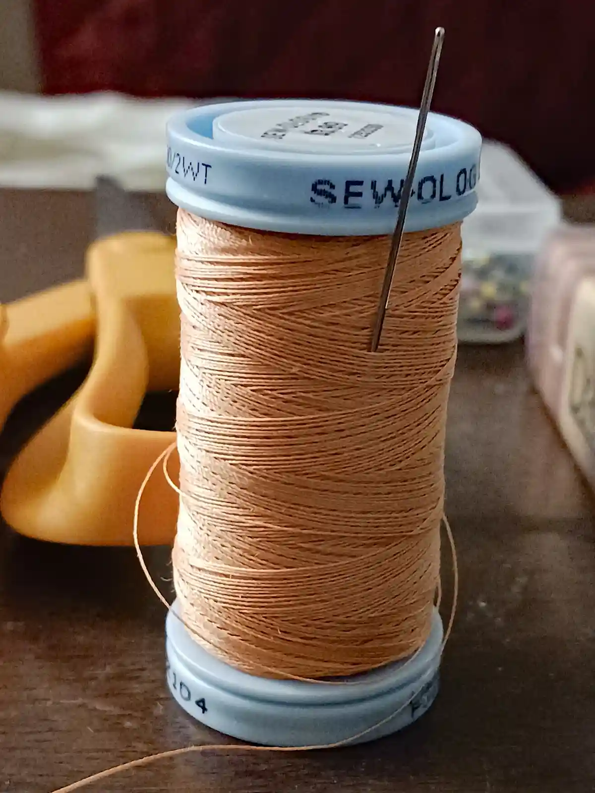 A close-up photo shows a spool of light brown sewing thread with a needle stuck vertically through its center. The spool has…
