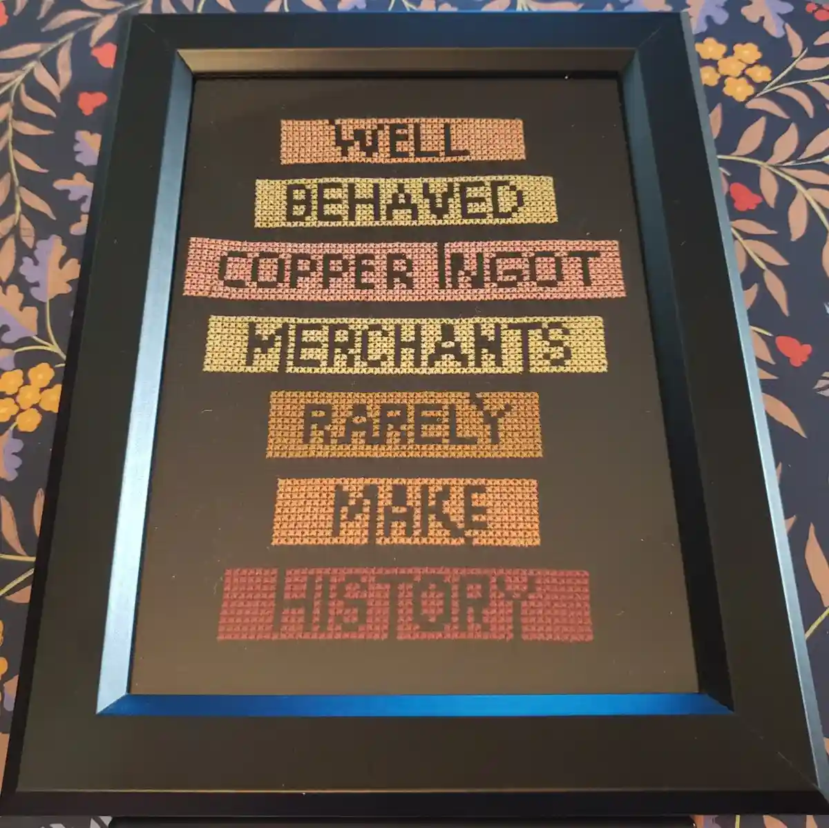 A framed cross-stitch artwork displays the phrase 'WELL BEHAVED COPPER NEGOTIATORS RARELY MAKE HISTORY' in blocky, pixelated…