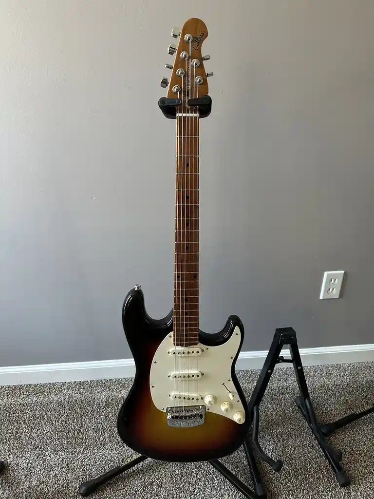A sunburst-finish electric guitar, likely a Squier Stratocaster, stands upright on a black guitar stand against a plain gray…