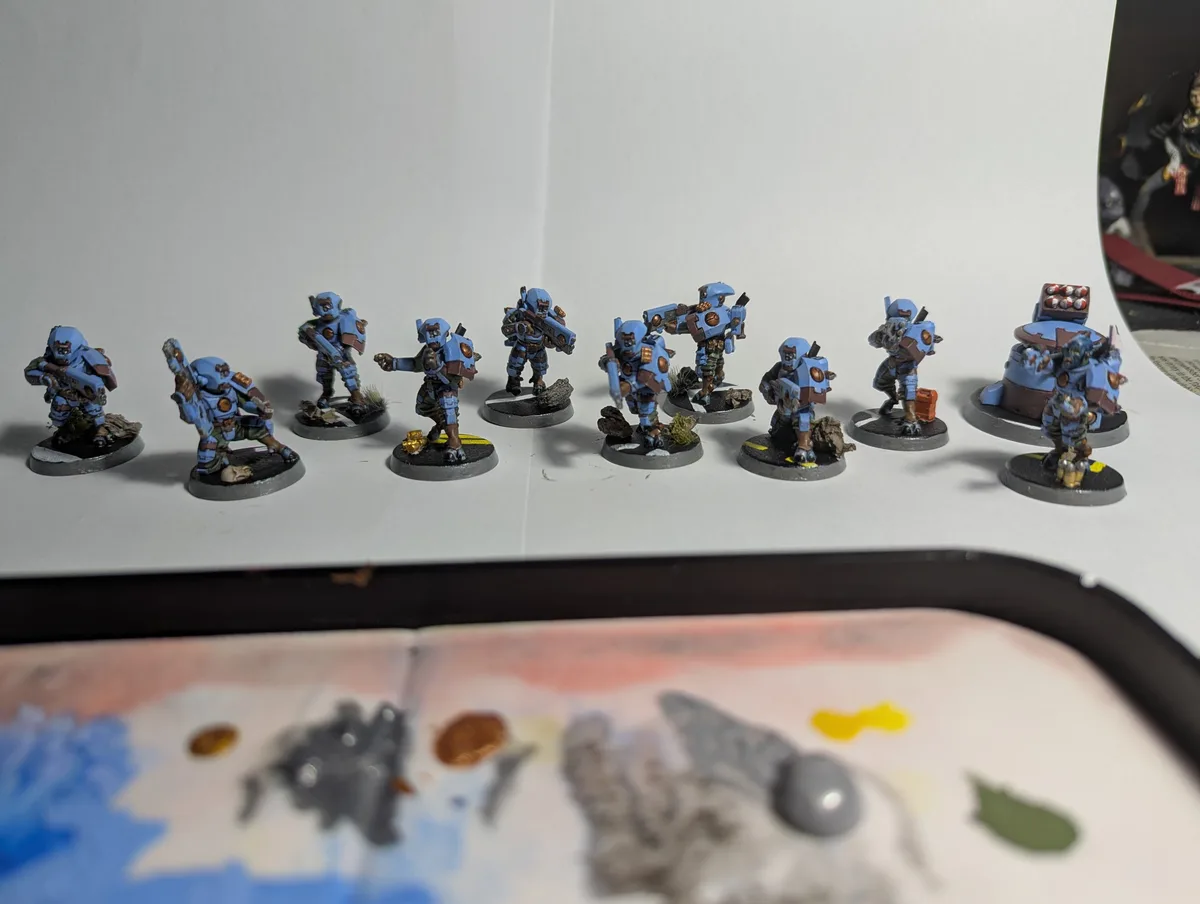 A row of painted miniature sci-fi soldiers, mostly in blue armor with some brown and gold accents, stands on a white…