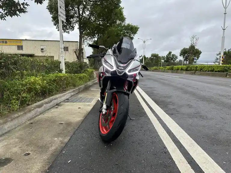 A silver and red Aprilia sport motorcycle is parked on the side of a wet asphalt road, facing the camera with its headlights…