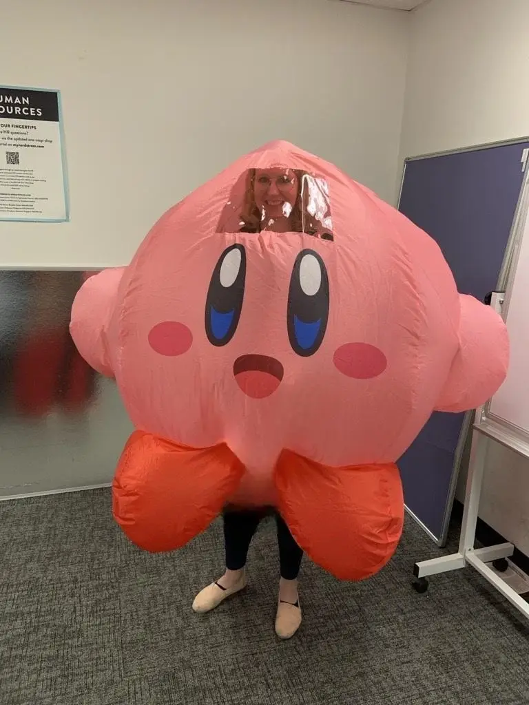 A person wearing a large, pink, inflatable Kirby costume stands in an office, with only their face visible through a clear…