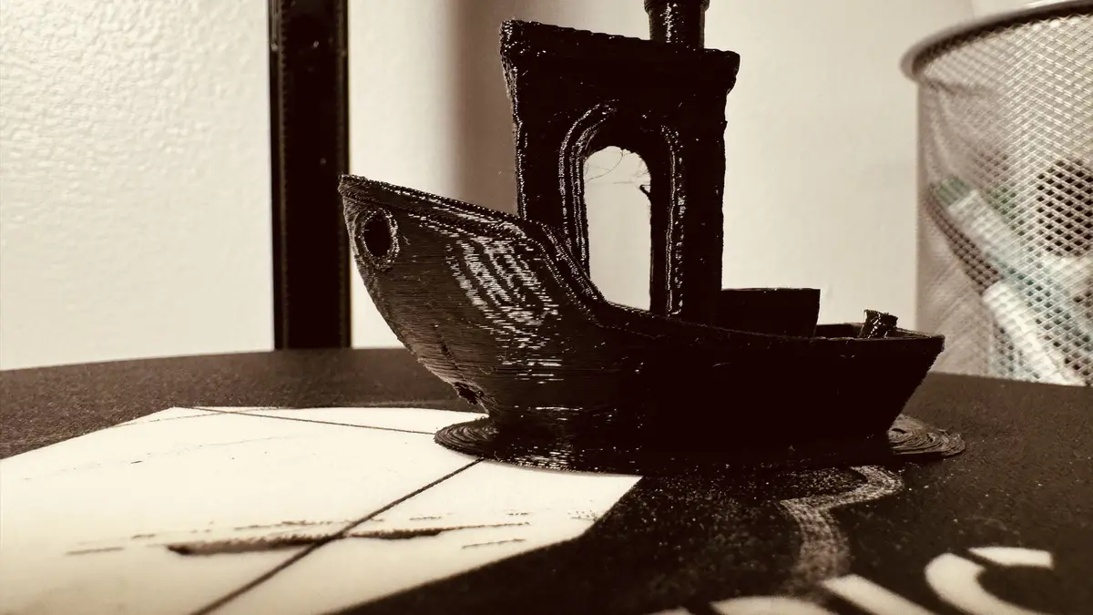 A small, black 3D-printed boat model sits on a tiled surface, its layered plastic construction visible. The boat has a…