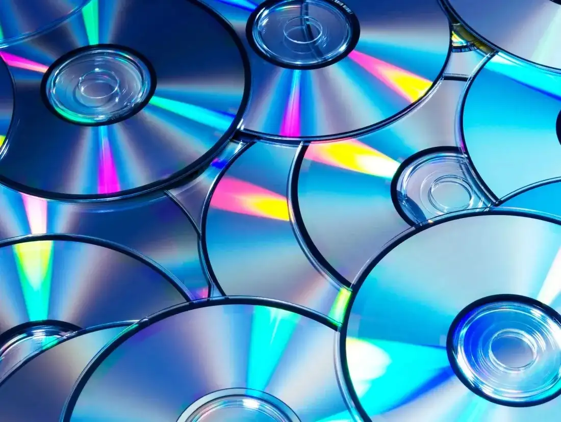 A close-up image shows numerous compact discs (CDs) overlapping and stacked, their reflective surfaces catching light to…