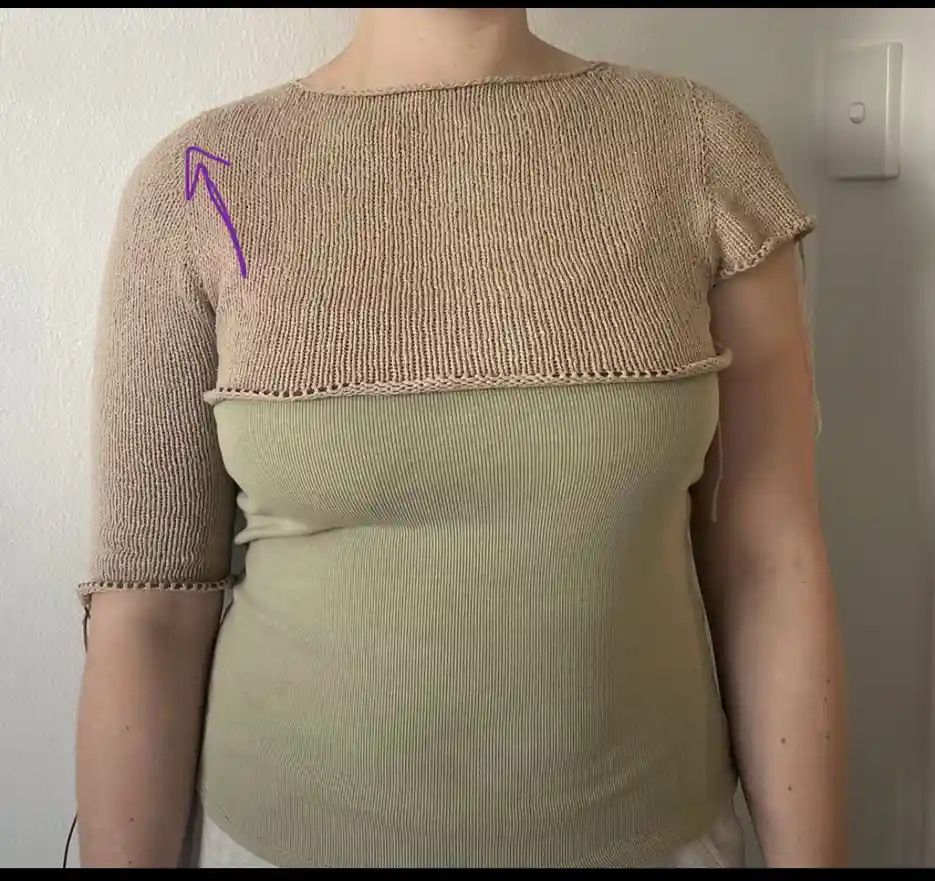 a person wears a beige knit top over a green ribbed tank top; a purple arrow points to the shoulder seam of the knit top…