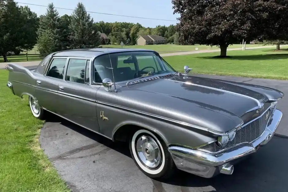 A shiny silver 1960s American sedan, likely a Chrysler or similar model, is parked on a paved driveway next to green grass…