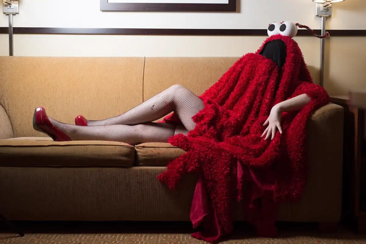A person lounging on a tan couch wearing fishnet stockings and red high heels, draped in a red, fuzzy Elmo costume that…