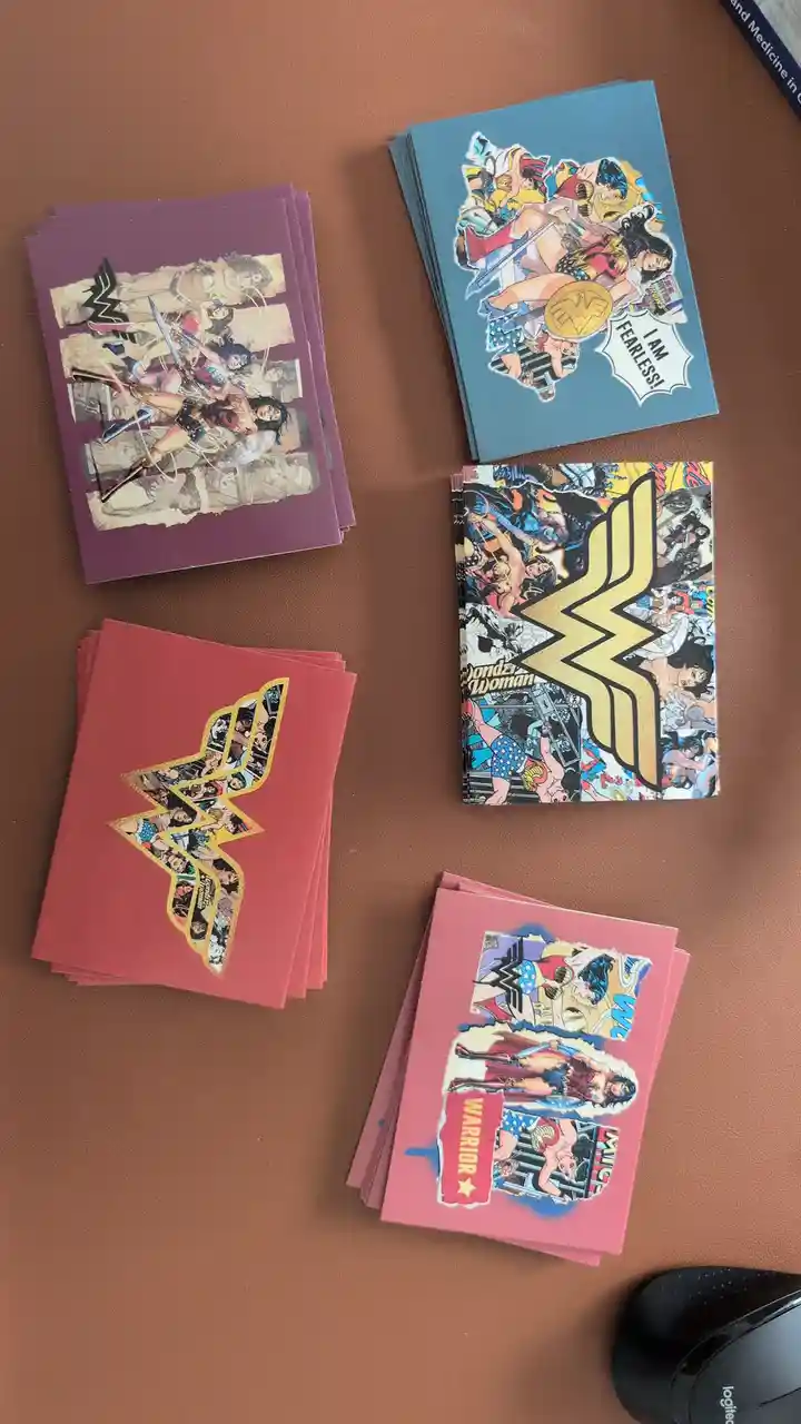 Five stacks of Wonder Woman themed notebooks or cards are arranged on a brown surface, each featuring different comic-style…