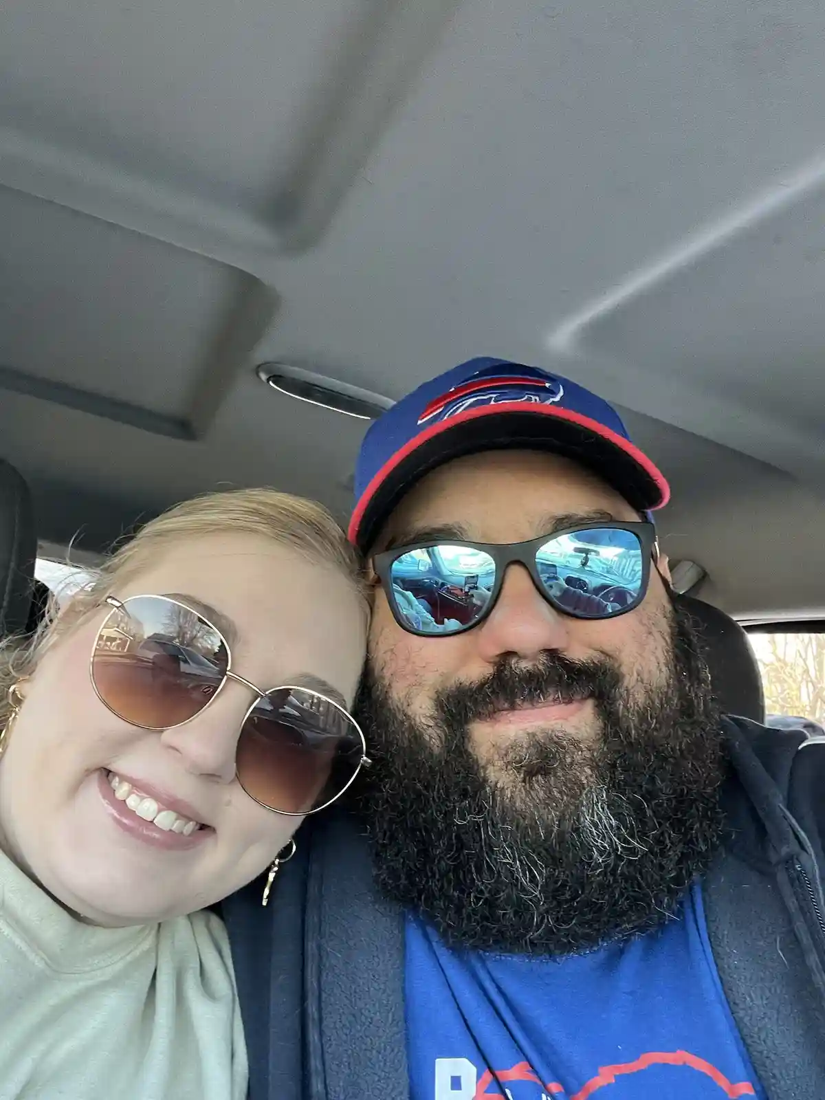 A selfie of a smiling couple inside a car, both wearing sunglasses. The man has a beard and wears a blue Buffalo Bills…