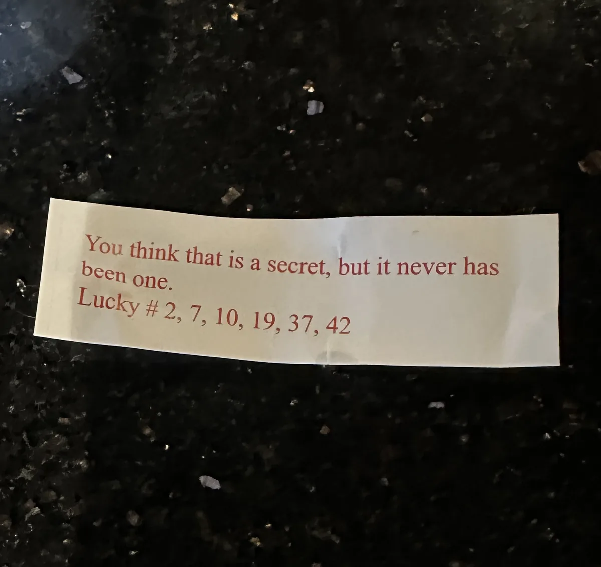 A crumpled white fortune cookie slip with red text lies on a dark, speckled countertop. The text reads: 'You think that is a…