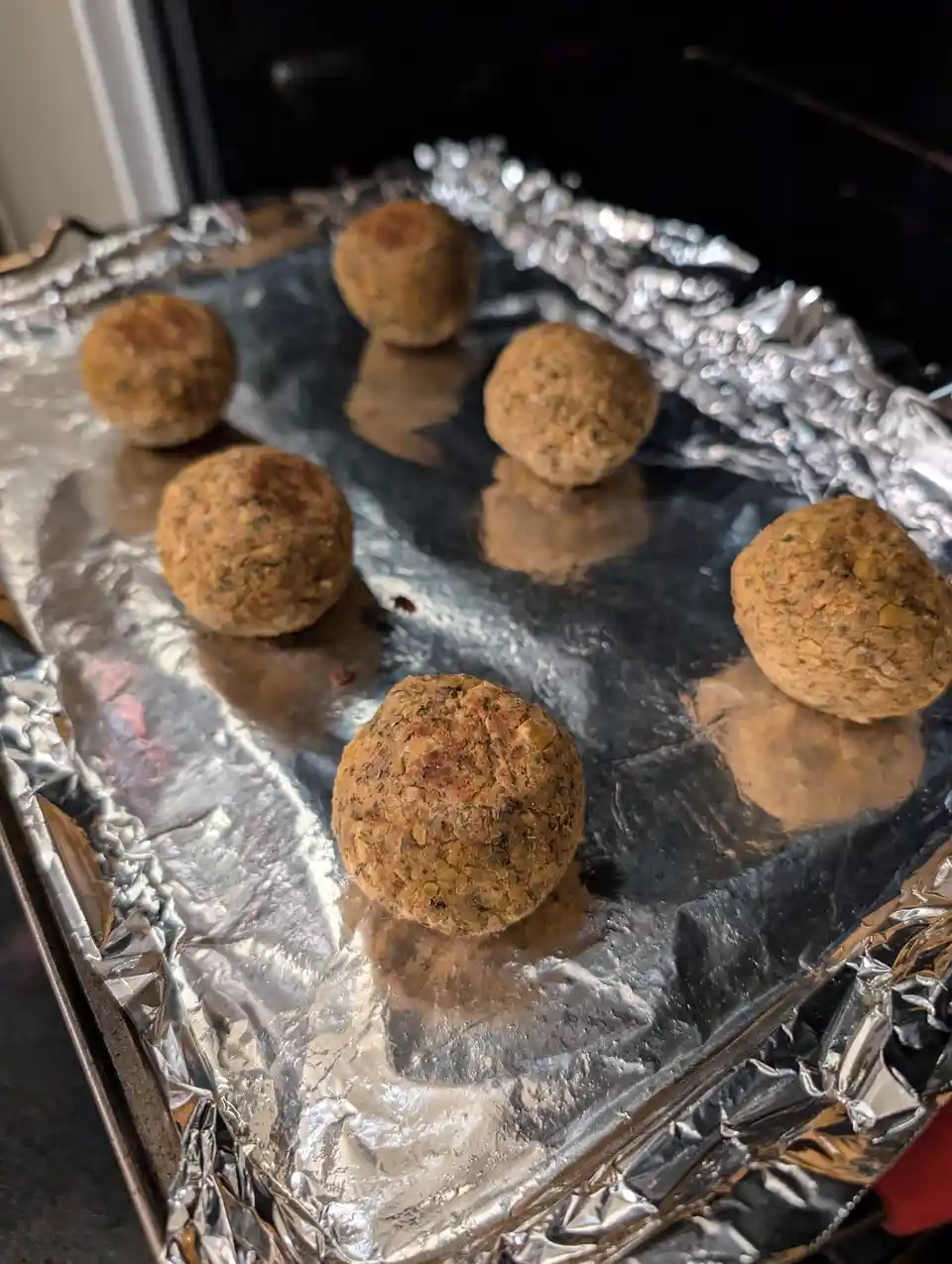 A close-up photo shows six golden-brown falafel balls resting on crinkled aluminum foil on a baking tray. The balls have a…