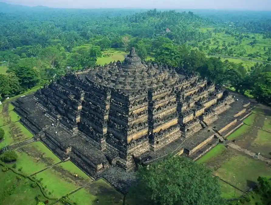 An aerial photograph shows the ancient, multi-tiered stone Borobudur temple in Indonesia, surrounded by lush green forests…