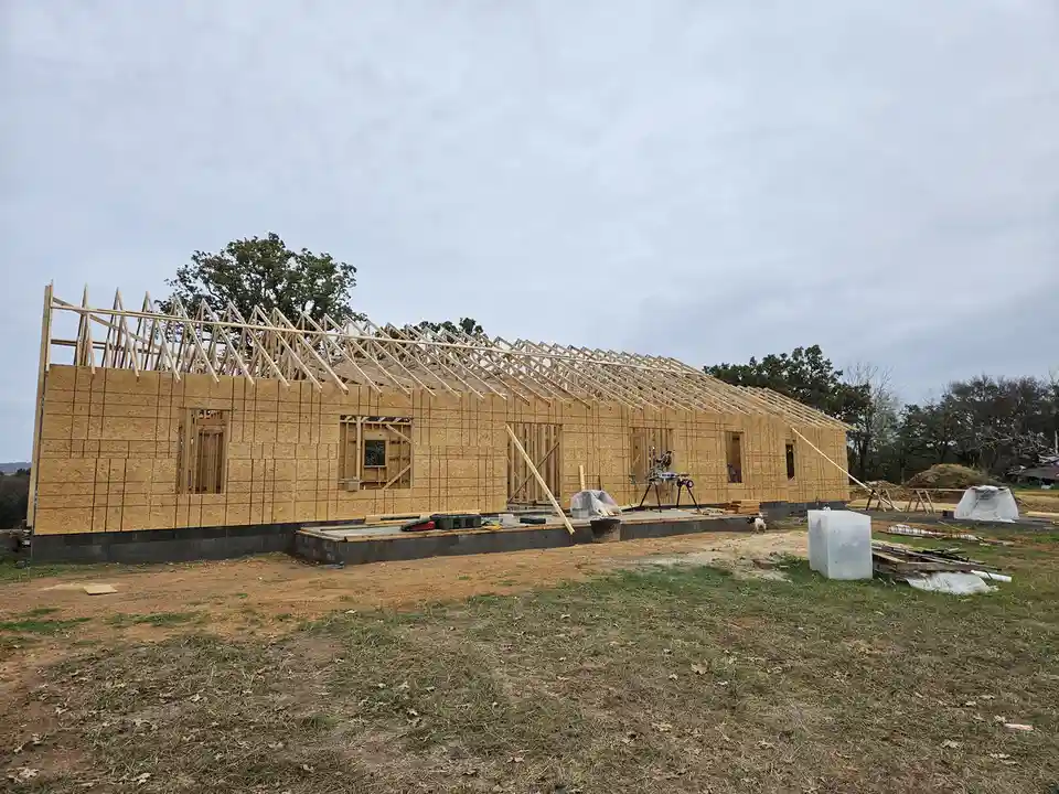 A house under construction with exposed wooden framing and OSB sheathing, surrounded by dirt and construction tools under an…