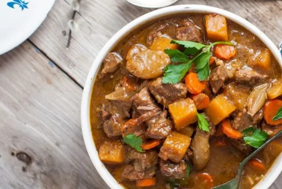 a close-up photo of a white bowl filled with hearty beef stew containing chunks of meat, carrots, potatoes, and parsley…