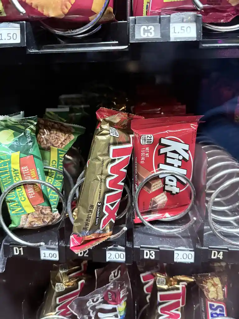 A close-up of a vending machine's snack selection showing a Twix bar and a KitKat bar side-by-side in adjacent slots, both…