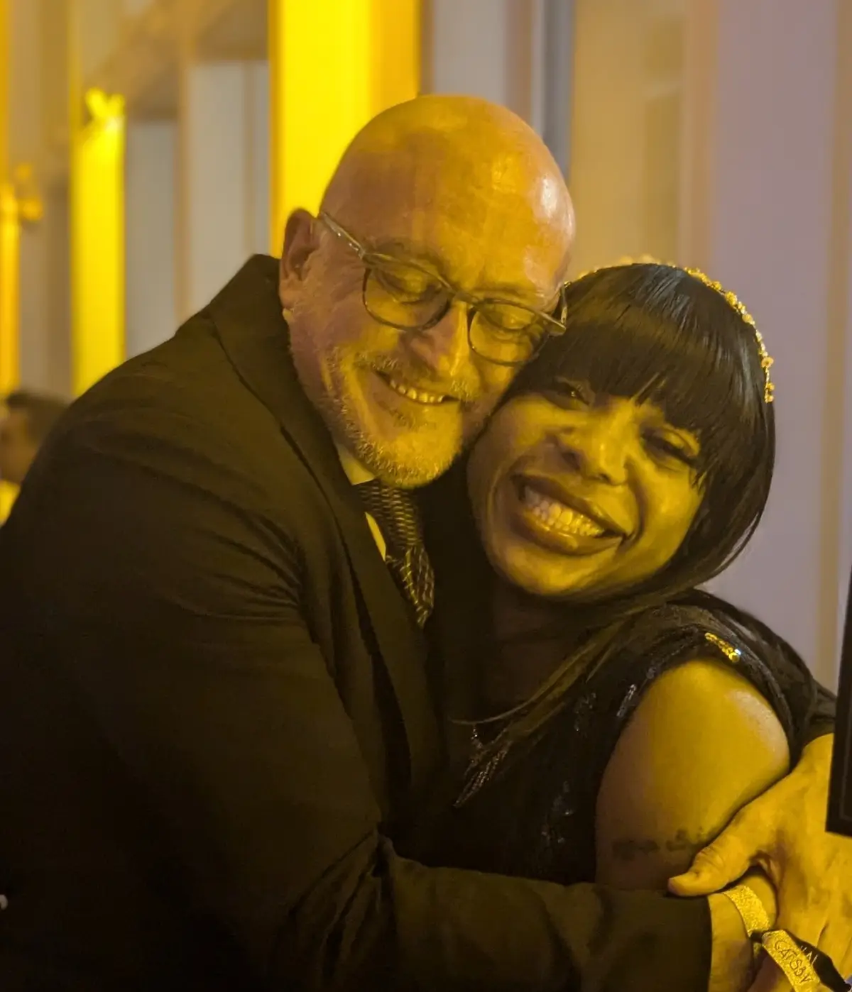 A bald man in glasses and a suit embraces a smiling woman in a black dress under warm yellow lighting. The overlaid text…