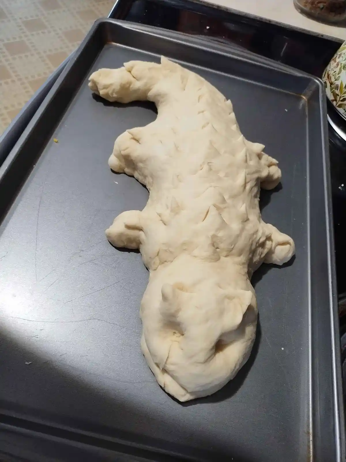 A pale, uncooked dough shaped like a crocodile with textured back and limbs, lying on a metal baking tray. The overlaid text…
