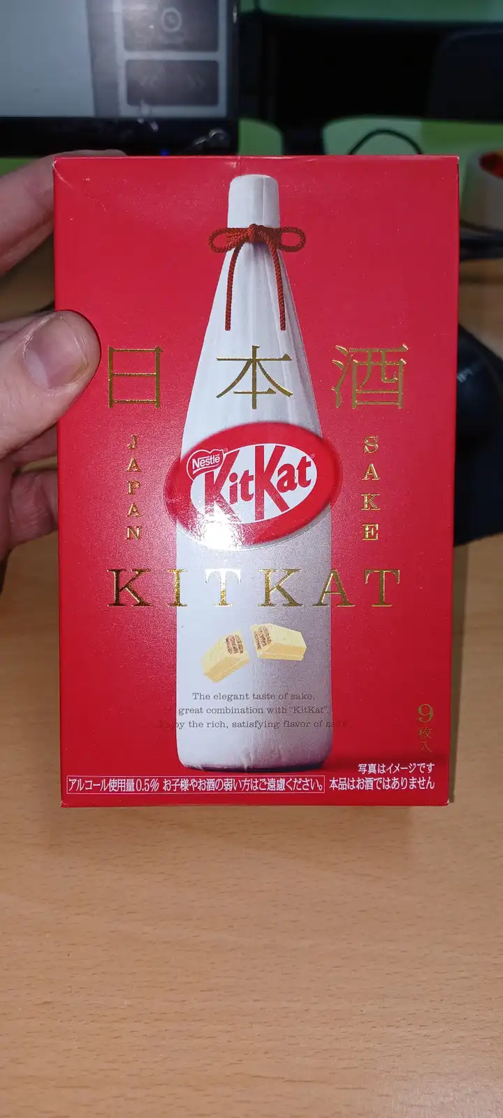 A hand holds a red KitKat box designed to look like a sake bottle, featuring Japanese text '日本酒' and English 'SAKE', with…