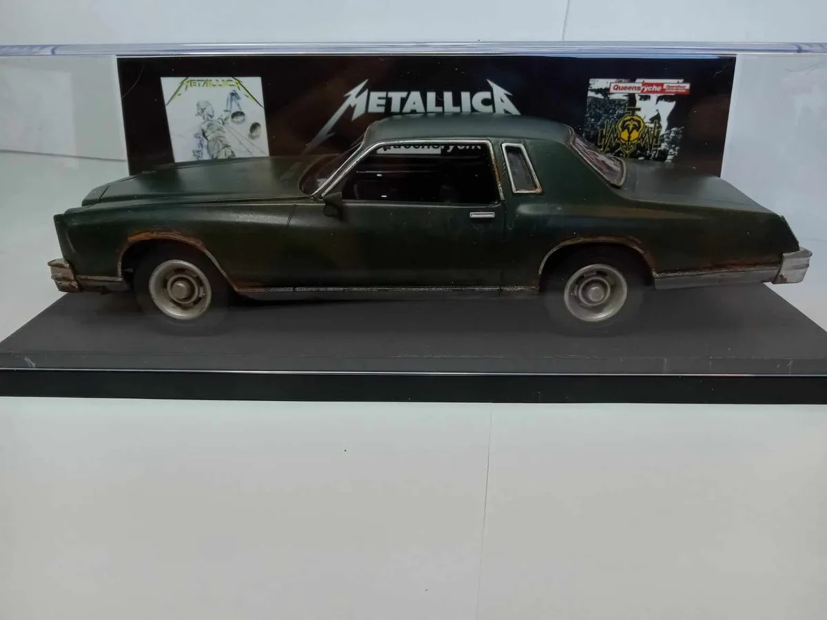 A green, rusty, and visibly worn model car is displayed in a clear case, with the band names 'Metallica' and 'Queensrÿche'…