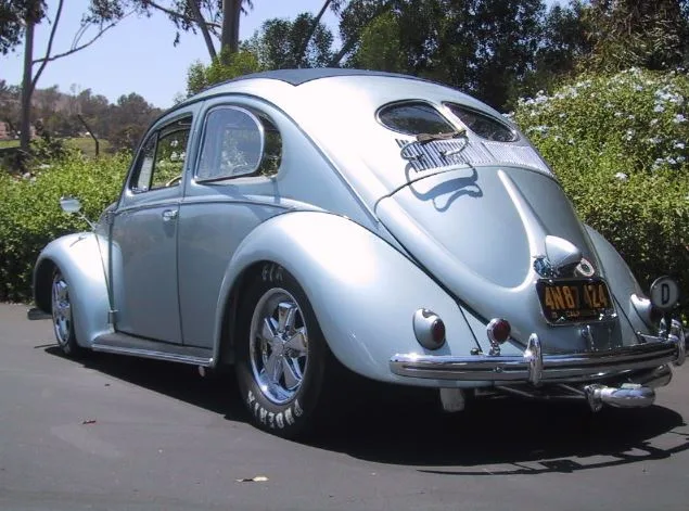 A silver classic Volkswagen Beetle is parked outdoors, viewed from the rear three-quarter angle. The car has custom chrome…