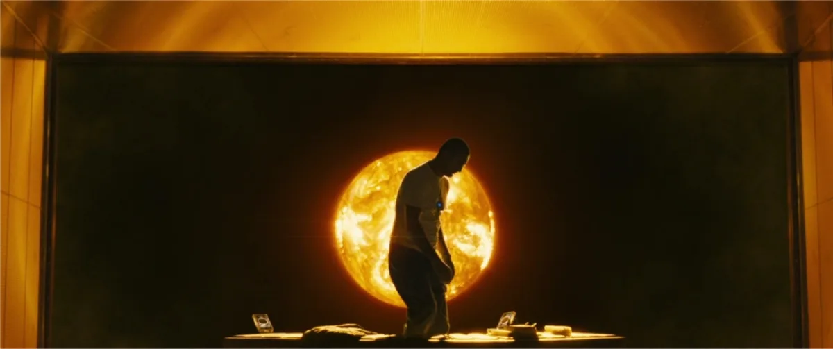 A man in a white t-shirt stands in front of a large screen displaying a fiery sun, looking downcast. The scene is framed by…