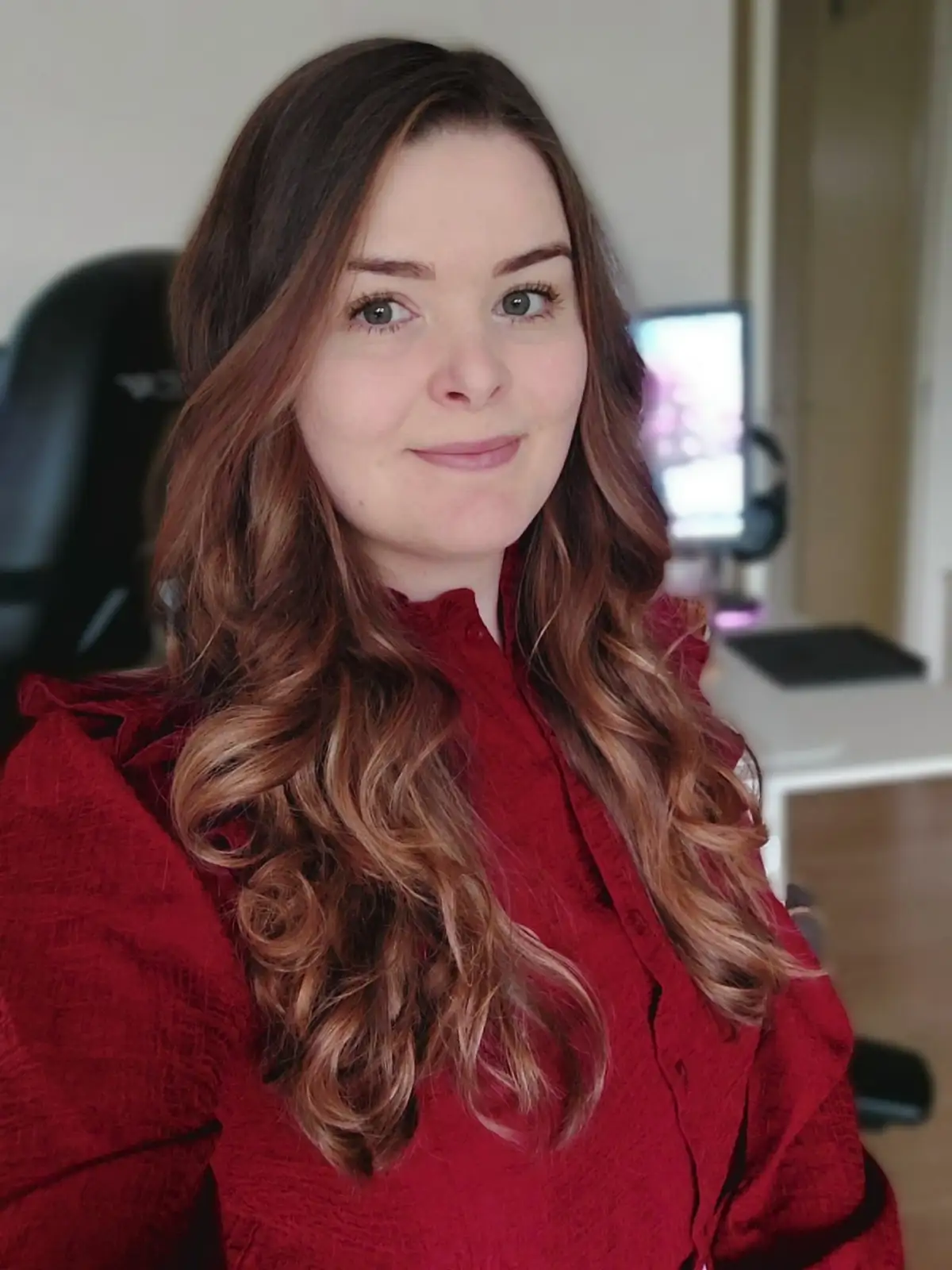 A woman with long, wavy brown hair and green eyes smiles at the camera while wearing a red blouse. Behind her, a black…