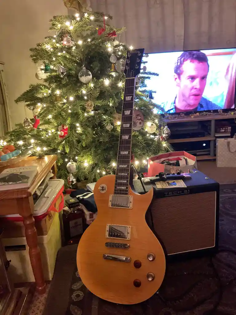 A light-wood electric guitar stands upright in front of a decorated Christmas tree with lights and ornaments, next to a Tech…