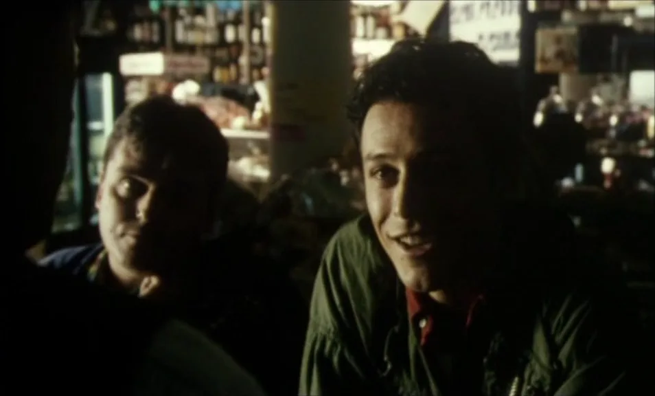 Two men are in a dimly lit bar or shop, one smiling broadly while the other looks unimpressed. The smiling man has dark hair…