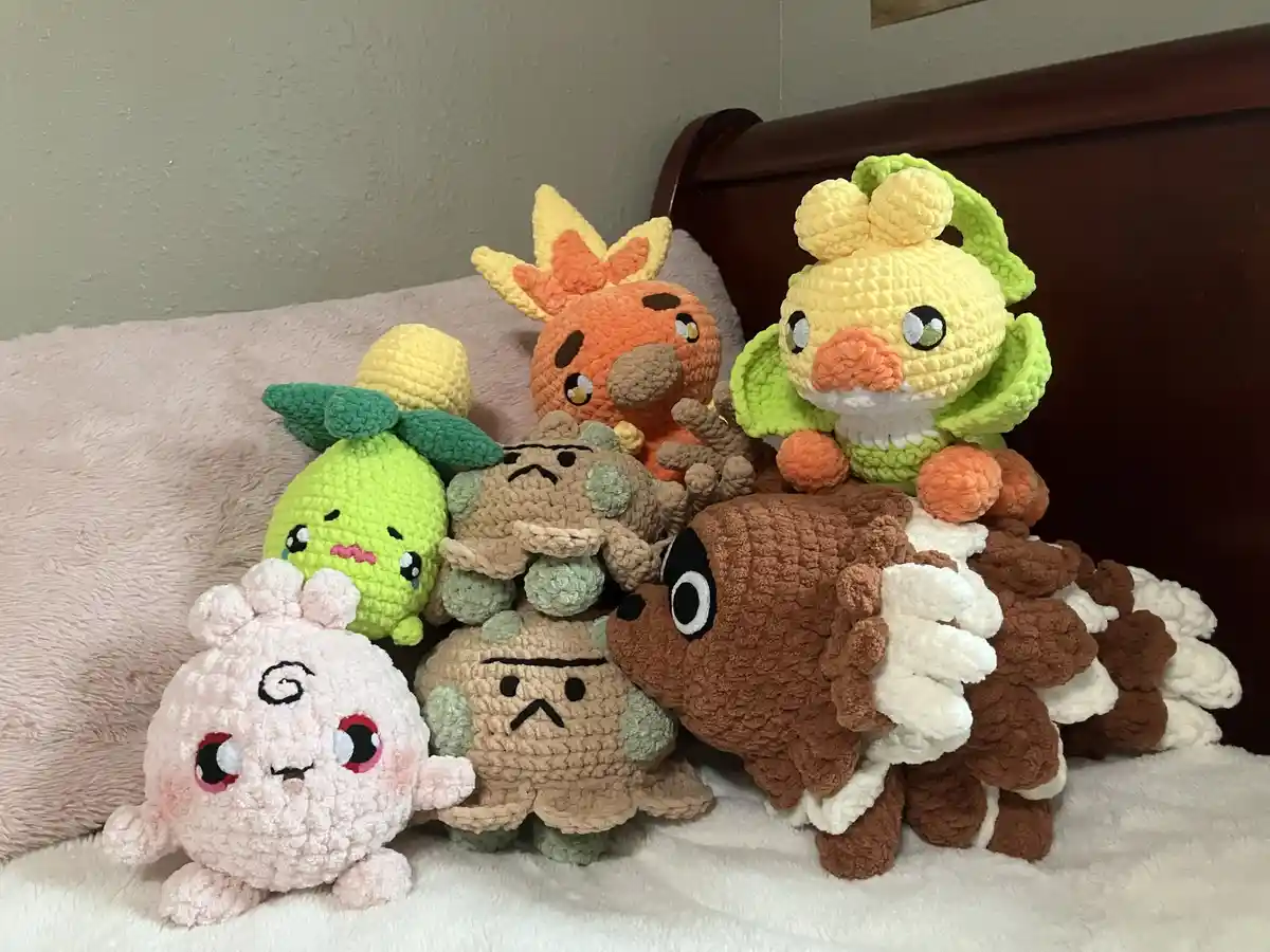 A collection of handmade, crocheted plush toys resembling Pokémon characters are piled together on a bed with a pink blanket…