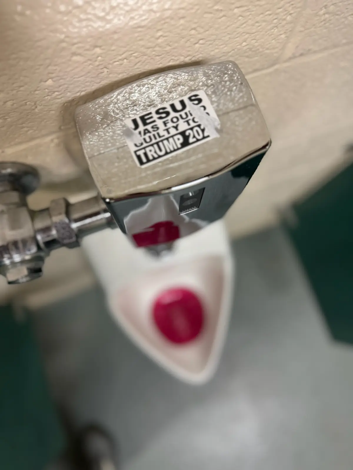 A close-up photo shows a white urinal with a chrome flush valve, viewed from above. A sticker on the valve reads 'JESUS WAS…