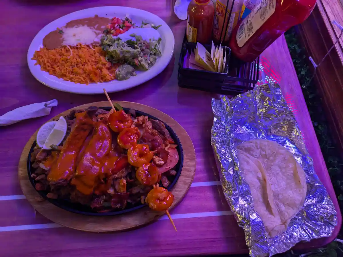 A table under purple lighting displays a large sizzling fajita platter with meat, shrimp, and sauce, alongside a plate of…