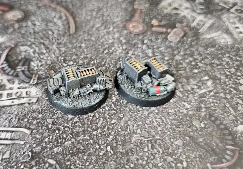 Two painted miniature tank models sit side-by-side on a gritty, debris-strewn surface. The tank on the left is intact and…
