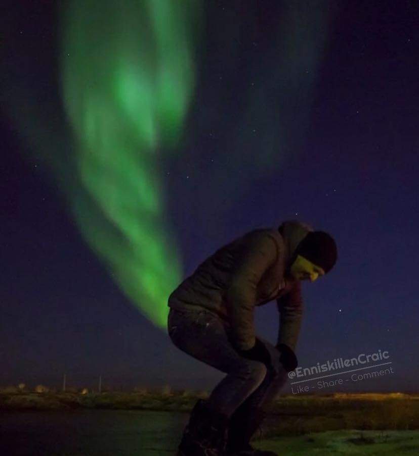 A person in winter clothing is bent over, appearing to cry, against a backdrop of vibrant green northern lights in a dark…