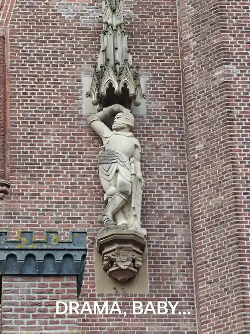 A stone statue of a medieval knight in full armor is mounted on a brick building, dramatically lifting his helmet off his…