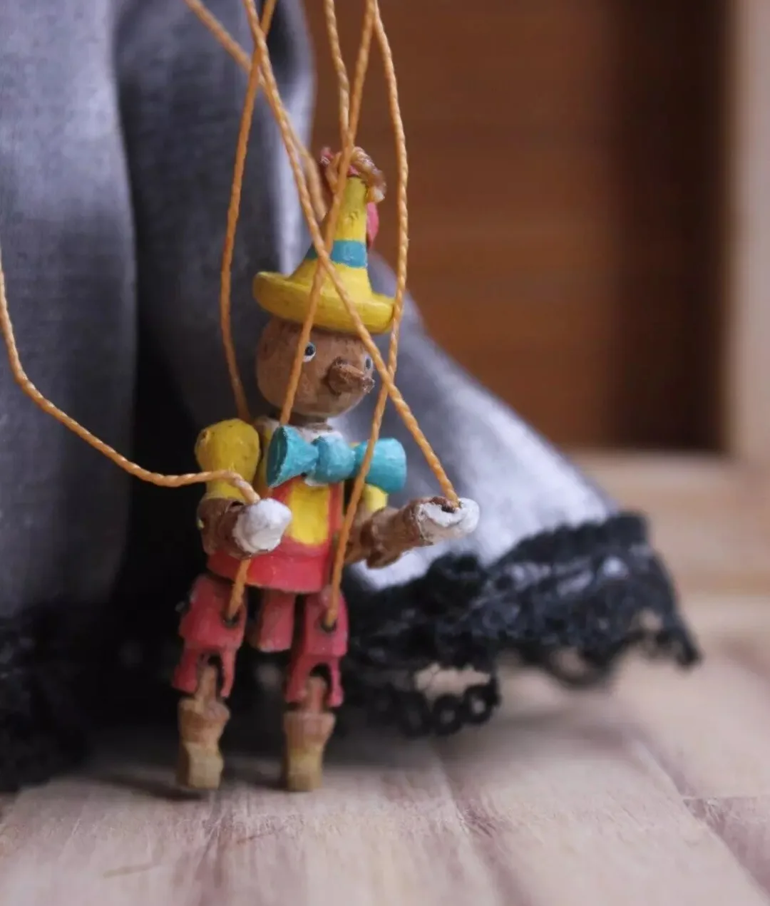 A close-up photo of a small, hand-painted wooden marionette puppet with a yellow hat, blue bowtie, and red pants, suspended…