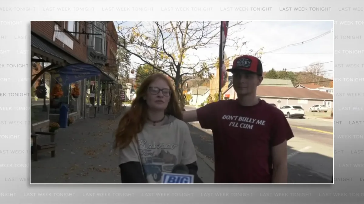 A young man wearing a red 'HOOEY' cap and a maroon t-shirt with the text 'DON'T BULLY ME I'LL CUM' stands with his arm…
