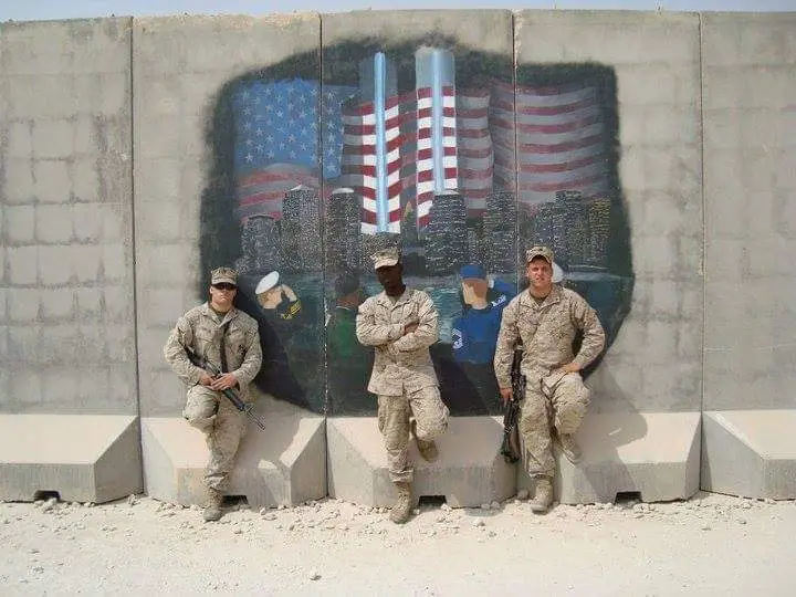 Four soldiers in camouflage uniforms sit or stand on concrete barriers in front of a large mural painted on a concrete wall…