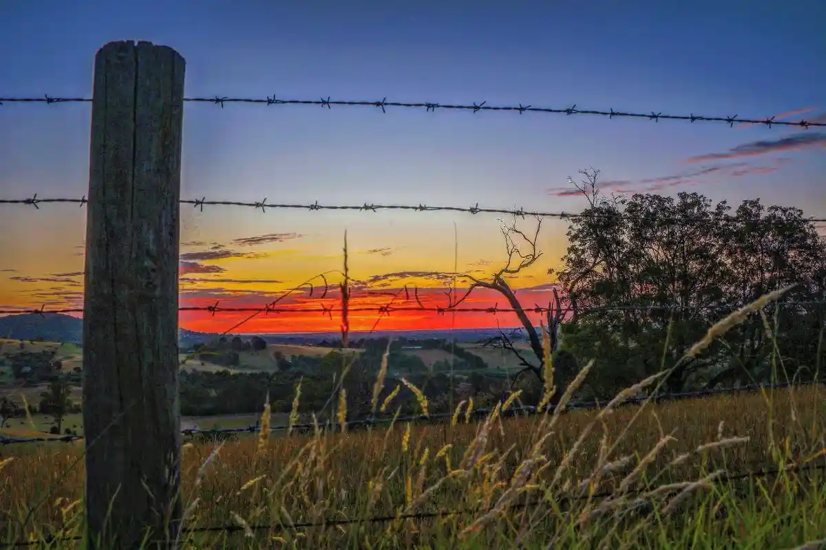 A scenic photograph shows a vibrant sunset over rolling hills, framed by a weathered wooden fence post and strands of barbed…
