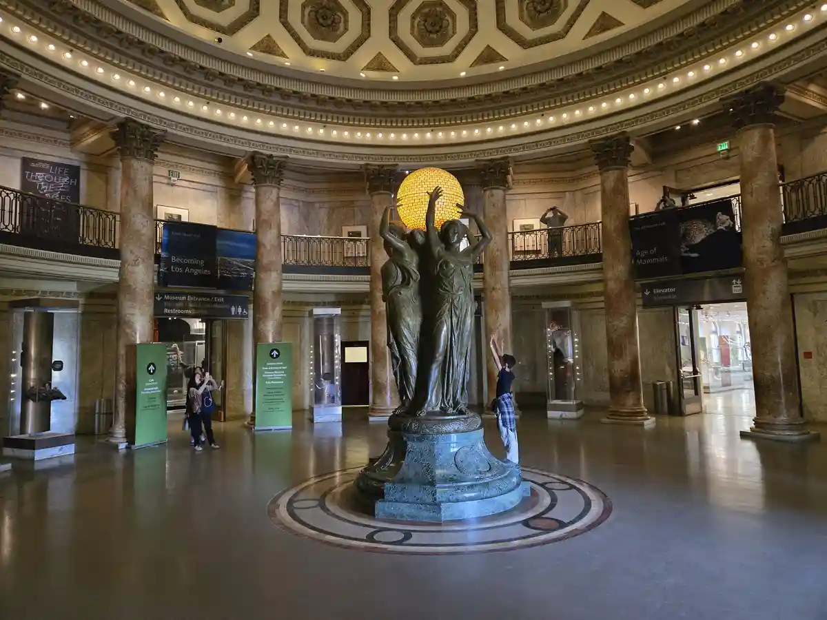 A wide shot of a grand museum rotunda with tall marble columns and a large central bronze statue of three figures holding up…
