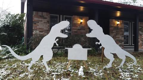 Two large white cutout T-Rex dinosaur figures stand facing each other in a snowy yard in front of a house, with a small…