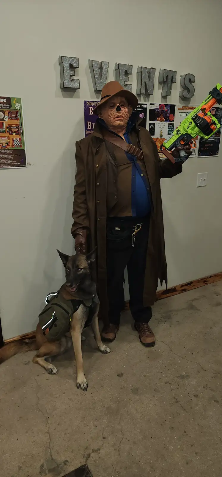 A person in a skull-faced, brown trench coat and fedora costume holds a bright green Nerf gun while standing next to a…