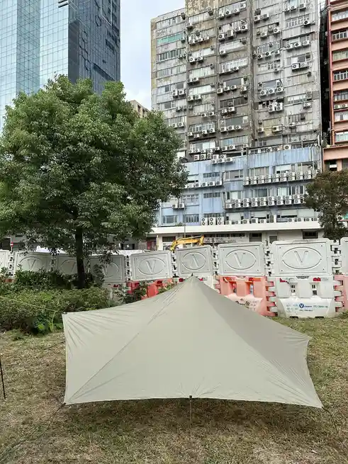 A beige tarp tent is set up on grass in front of construction barriers with 'V' logos, with a backdrop of a weathered…