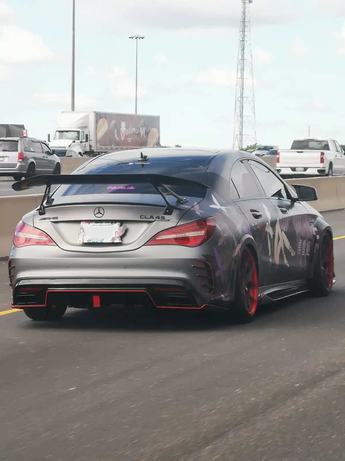 A heavily modified silver Mercedes-AMG CLA45 sports car with a large rear wing, red accents, and anime-style decals is…